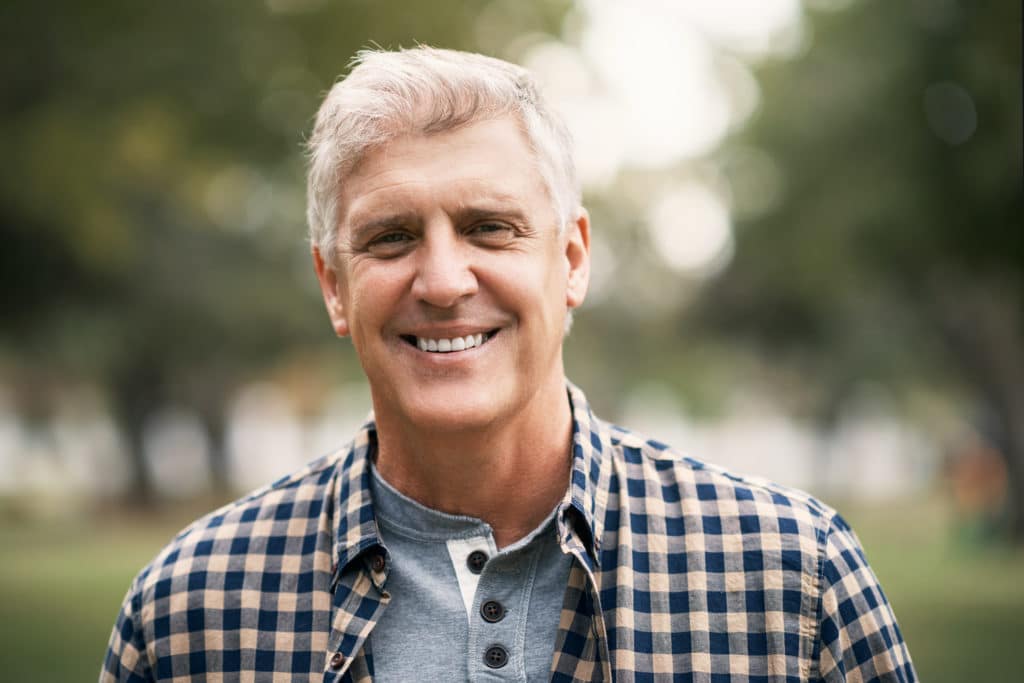 A happy mature man smiling with a stripped button shirt on in a park, the background is blurred.