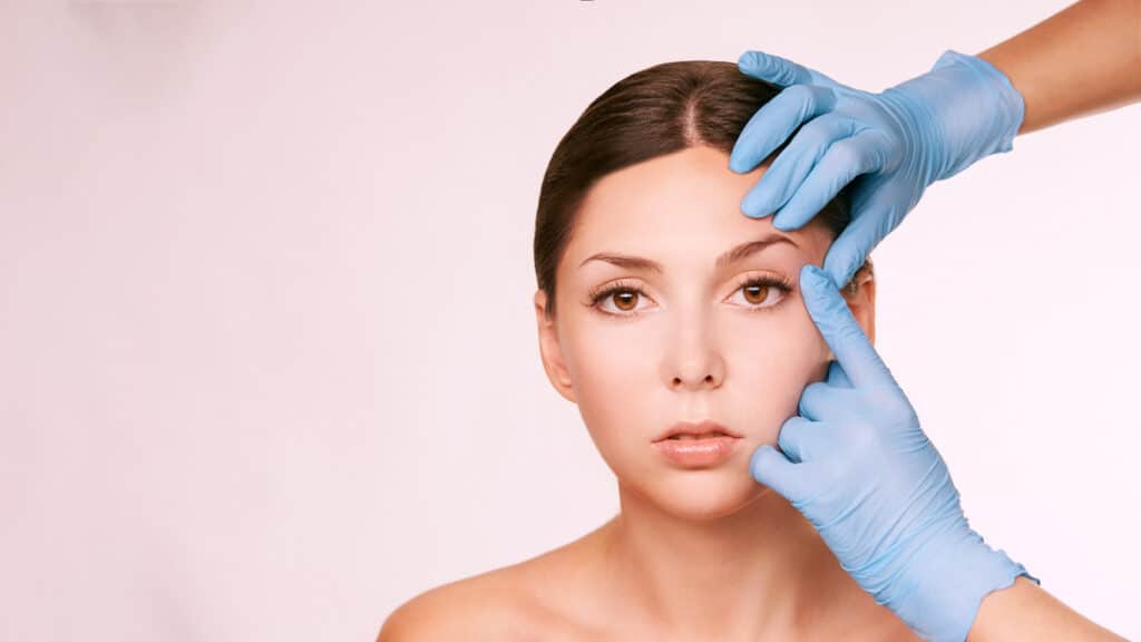 headshot of woman sitting while doctor inspects her eye against grey background