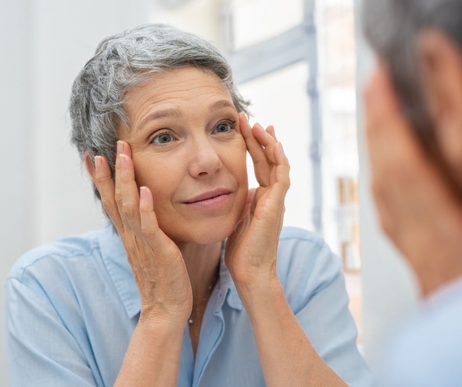 Beautiful senior woman checking her face skin and looking for blemishes. Portrait of mature woman massaging her face while checking wrinkled eyes in the mirror.