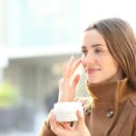 Satisfied woman applying moisturizer cream on her face in winter in the street