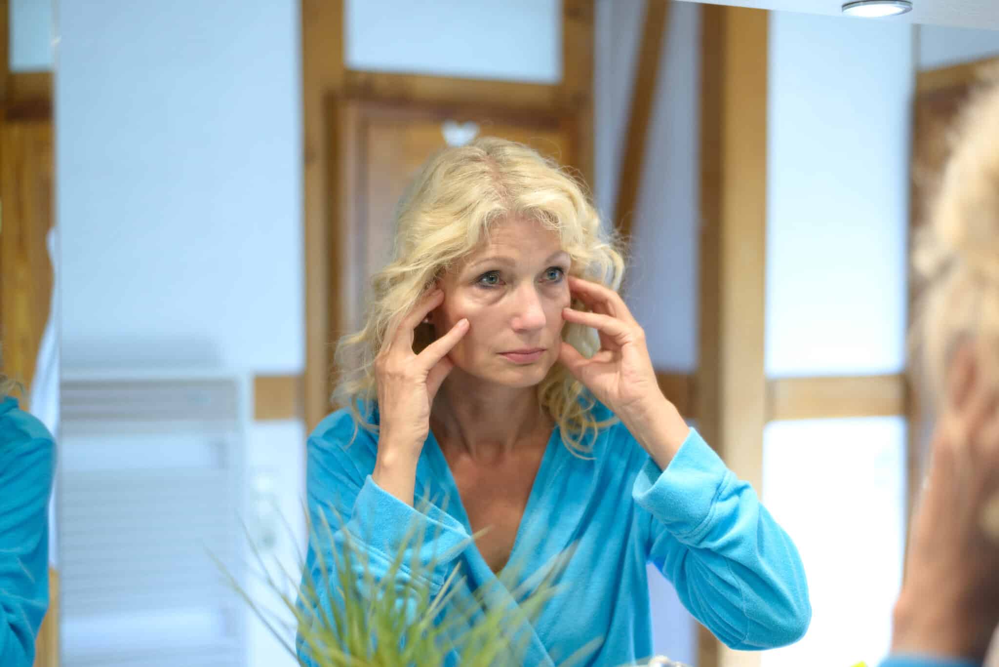 A woman looking in the mirror considering lower eyelid blepharoplasty vs. dermal fillers